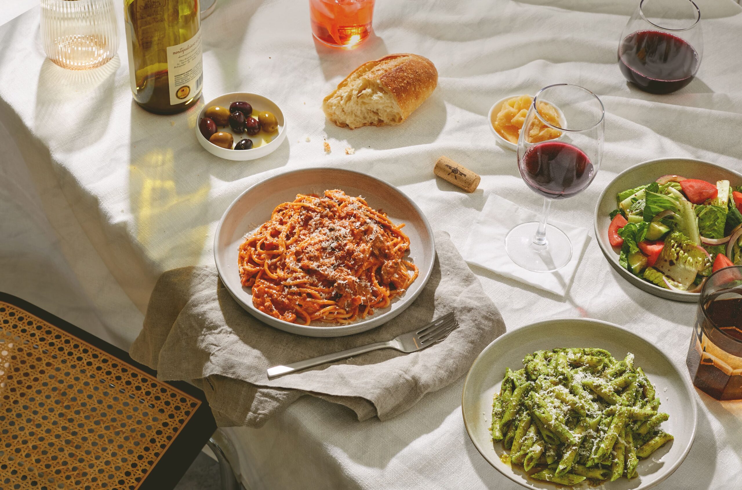 Image of salad, spaghetti marinara, and pesto penne on a table with wine and bread accents