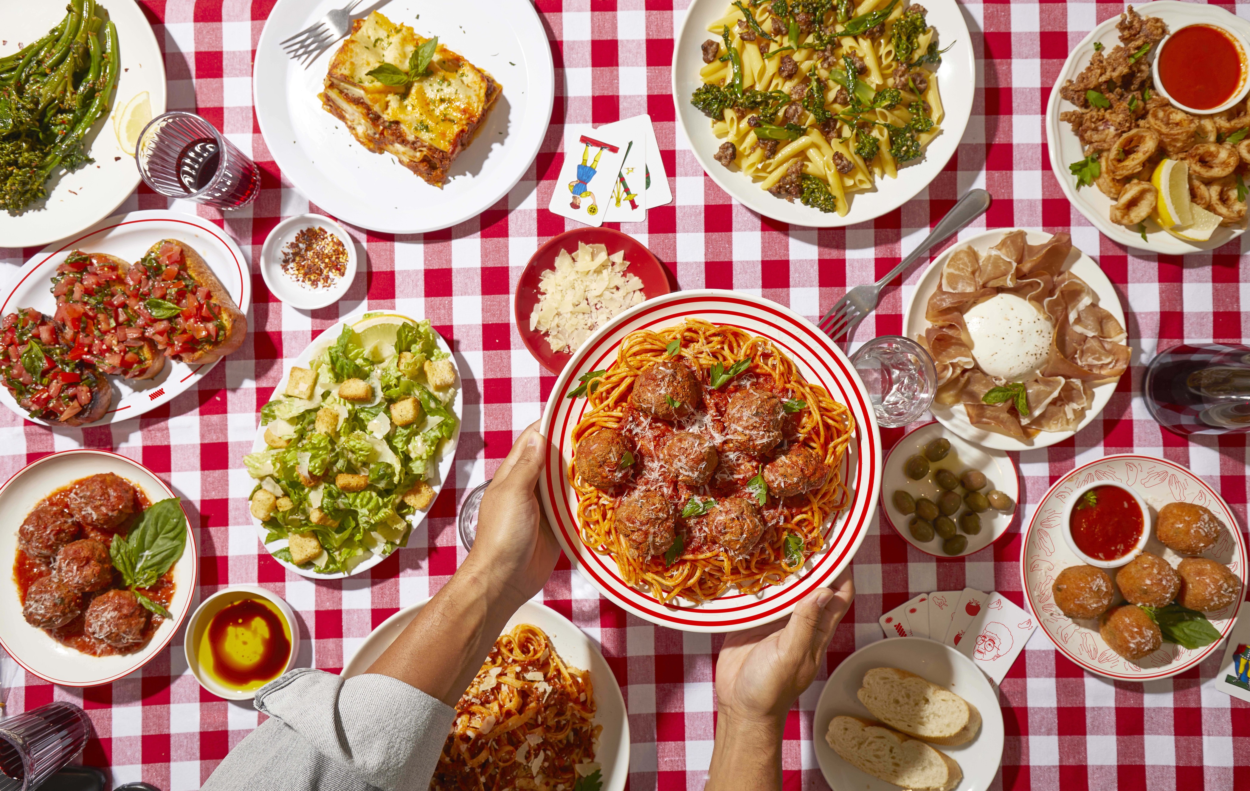 Image of various pastas, bruschetta, meatballs, salads, prosciutto, and wine on a red and white traditional table cloth
